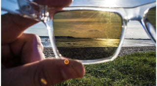 A hand holds clear sunglasses against a sunny beach, demonstrating the polarized sunglasses difference.