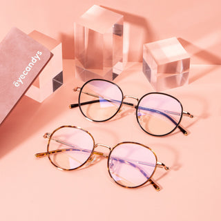 Two pairs of round eyeglasses with gold and black frames are displayed on a pink surface, alongside clear glass blocks and a pink Eyecandys case. The lenses have a slight purple-blue reflection.