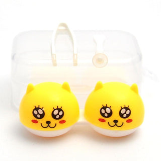 Two yellow, cat-shaped contact lens cases with cute cartoon faces. A transparent plastic case with tweezers and a suction tool is in the background.