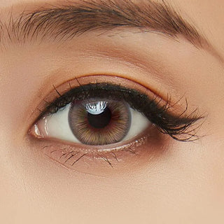 A close-up of a human eye with long lashes and a polished brow, featuring a light brown iris enhanced by Acuvue Define Fresh Grayzel (30pk) contacts from Acuvue. The eyelid shows subtle, natural makeup for a refined look.
