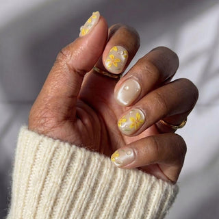 A hand wears EyeCandys' After Rain Press-On Nails, with yellow floral designs and white pearls on a nude base. Paired with a cream knit sweater and gold rings, sunlight highlights the reusable nails’ elegant look.