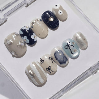 The All Tied Up Press-On Nails by EyeCandys feature handmade designsโflowers, abstract patterns, metallic accents, and zodiac-style symbolsโin beige, blue, white, and silver, all displayed in a clear case.