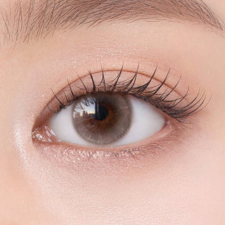 Close-up shot of model's eye adorned with Chuu Aube Pie Moon Beige (10pk) color contact lenses with prescription, complemented by minimalist eye makeup, showing the brightening and enlarging effect of the circle contact lens on dark brown eyes.