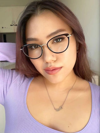 Wearing the Aura Cat Eye glasses by EyeCandys, a woman with long brown hair and glasses looks directly at the camera, smiling slightly. She wears a light purple top and a "cutie" necklace in a bright indoor setting.