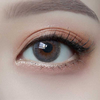 A close-up of a human eye with brown irises and long, curled lashes wearing Molak 1-Day Bubble Blue (10pk) lenses by Molak. Peach-toned eye shadow colors the eyelid, while a window’s reflection glimmers in the eye.