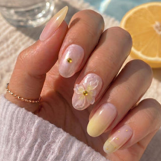 A hand with long, almond-shaped Daisy Does It Press-On Nails by EyeCandys showcases a pastel ombre salon gel look with gold studs, 3D florals, and delicate gold dots. Background: lemon slice and textured white surface.