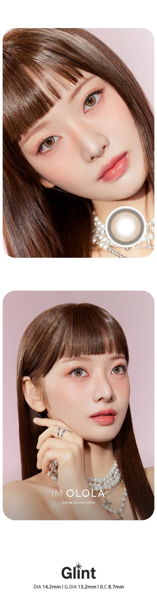 A woman with straight brown hair and bangs wears Olola Glint 1-Day Grey (10pk) color contacts by Olola, pearl jewelry, and soft pink makeup, posing against a pale pink background. "Glint" is displayed at the bottom.