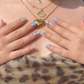 Hand wearing reusable Gilded Cirrus press on nails with impressionist sky blue cloud art, gold celestial details, and elegant short oval shape.