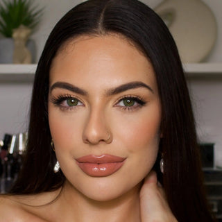 A woman with long, straight dark hair and clear, greenish eyes, accentuated by EyeCandys Isla Emerald Green non-prescription contact lenses from EyeCandys, is looking at the camera. She has flawless makeup, a nose ring, and is holding her right hand to her neck. The background appears to be a blurred room with some decor and objects.