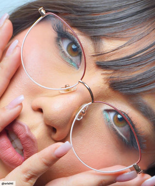 A close-up of a person wearing EyeCandys' Madame Cat Eye glasses with bold teal eye makeup and natural lipstick. Their manicured hands frame their face, drawing attention to the stylish cat-eye frames.