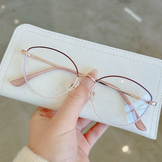 A hand holds the EyeCandys Madame Cat Eye glasses—round, pink-rimmed cat-eye frames with clear lenses—resting on a white case. Lightweight metal adds a modern twist to this classic style.