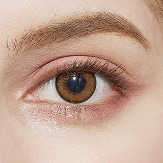 A close-up of a person's eye wearing NEO Vision Cosmo Brown circle lenses by NEO Vision, complemented by natural-looking makeup and a well-groomed eyebrow.