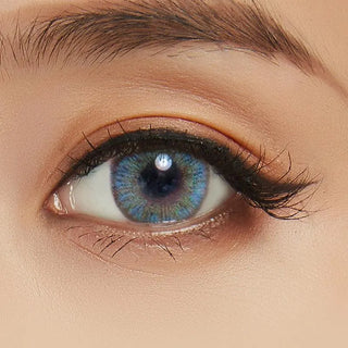 A close-up shows an eye wearing NEO Vision Cosmo Blue lenses by NEO Vision, with blue and green patterns. The eye is framed by groomed brows, dark lashes, and softly blended nude eyeshadow.