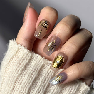 Hand wearing EyeCandys Nyx press on nails with translucent taupe lavender cat eye base and gold celestial sun and star nail art.