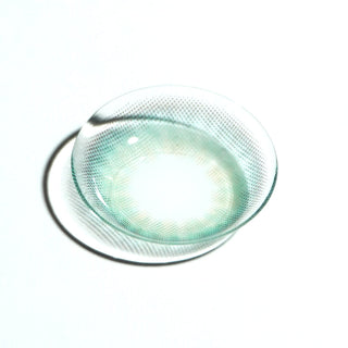 A single EyeCandys Pink Label Mallorca Blue contact lens, featuring a translucent blue with subtle green tint and dotted pattern, rests on a white surface and casts a soft shadow.