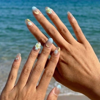 Hand wearing EyeCandys Lei’d Back press on nails featuring nude to blue ombre gradient, gold shimmer, and 3D plumeria flower accents.