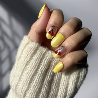 Hand showcasing EyeCandys Cherry Picnic press on nails with soft pastel yellow chrome finish and cherry detailing.