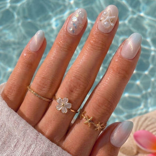A hand displays EyeCandys' Pearl Jam Press-On Nails—pastel ombre with white floral, pearl, and glitter accents—adorned with gold flower and starfish rings against a blue pool background.