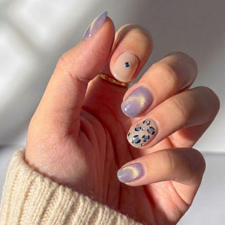 A manicured hand wears EyeCandys Purr-ple Reign Press-On Nails: pastel purple cat eye nails with shimmer, plus nude accent nails—one with a blue gem and one with black and blue leopard print. A cream sweater completes the look.