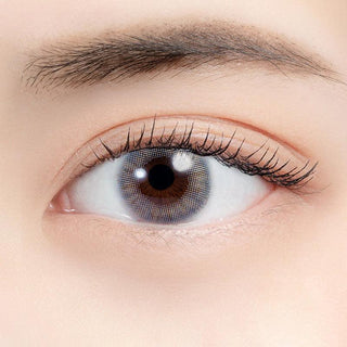 A close-up of a human eye wearing N's Collection Tama Konnyaku Grey (10pk) contacts shows fine eyelashes and a defined eyebrow above. The smooth, even-toned skin highlights the vibrant effect of these grey coloured lenses from N's Collection.