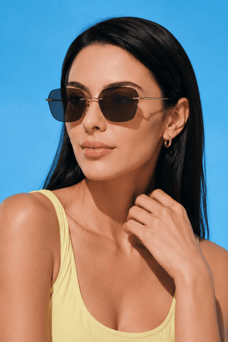 A woman with long dark hair wearing EyeCandys Dusk Rimless Rectangular glasses, a yellow sleeveless top, and a small hoop earring poses against a bright blue background, lightly touching her neck.