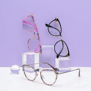 Three pairs of EyeCandys Aura Cat Eye glasses in pink, black, and gray are shown on clear acrylic blocks with a small transparent sphere nearby, all set against a light purple background.
