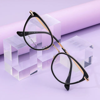 A pair of EyeCandys Aura Cat Eye eyeglasses in black and gold sits on clear acrylic blocks against a pastel purple backdrop, highlighting the elegance and wide fit of this stylish frame.
