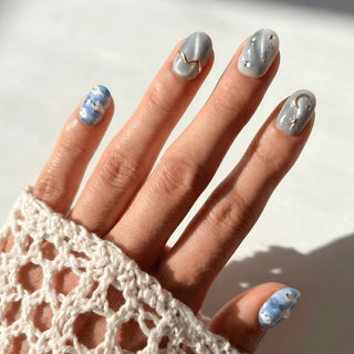 Hand displaying EyeCandys Gilded Cirrus press on nails with ethereal sky blue cloud design, metallic moon and star accents, and soft cat eye shimmer.