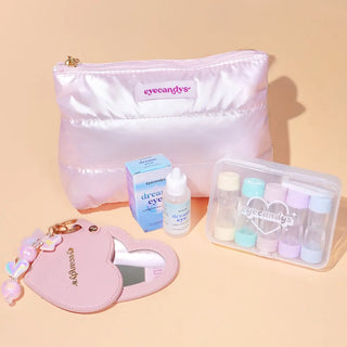 A pastel pink EyeCandys travel kit featuring a shiny puffy makeup pouch, a blue packaging box and bottle of Dream Eye lubricant eye drops, a clear case with colorful contact lens containers, and a pink heart-shaped mirror keychain with charms.