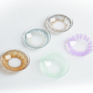 Macro shot of assorted colored contact lenses (brown, blue-grey, grey, green, purple) showing a variety of limbal ring designs (dashed, solid, graduated) and cosmetic patterns on a light background