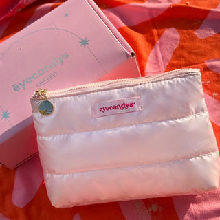 A soft, pink puffy makeup pouch with a quilted texture and a gold zipper, featuring a round charm and a stitched "eyecandys" label in pink. It’s placed on a vibrant orange and pink fabric, next to a pastel pink EyeCandys box.