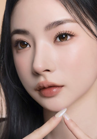 A female model with natural dark eyes wearing i-Sha Reborn Oak Haze contact lenses, complemented by subtle eyeshadow and peach lipstick