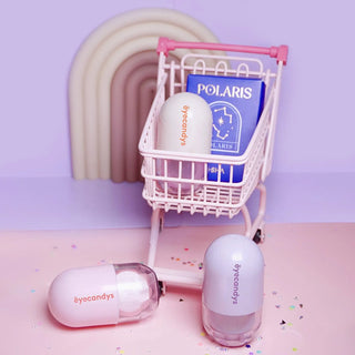 A small white shopping cart, partially tipping over, contains a blue "Polaris" booklet, a contact lens case, and two pastel-colored capsule containers labeled "EyeCandys Pill-Shaped Contact Lens Case Kit." Two more EyeCandys Pill-Shaped Contact Lens Case Kits, one pink and one purple, are on the pink surface next to the cart against a light purple background.
