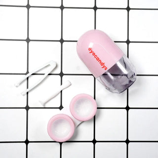 A Pill-Shaped Contact Lens Case Kit on a white and black grid background. The kit includes a pink and transparent contact lens case with the "EyeCandys" logo, soft-tipped tweezers, and a lens applicator stick. The case is opened displaying two compartments for the lenses.