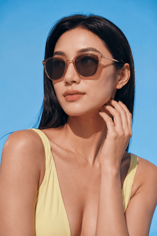 A woman in a yellow swimsuit poses against a bright blue sky, wearing EyeCandys Posh Square full-frame glasses, touching her face and gazing into the distance with a relaxed expression.