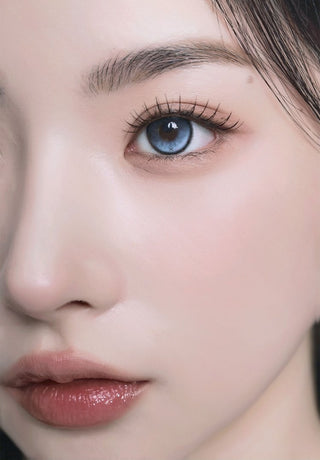 A female model with natural dark eyes wearing i-Sha Reborn Reborn Sky Veil contact lenses, complemented by subtle eyeshadow and peach lipstick