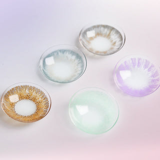Macro shot of various color contact lenses in shades of grey, brown, blue, green and purple, with various graphic diameters and limbal ring designs, on a light purple background