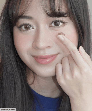 Female model trying on the Sunlit Hazel circle contact lens in one eye, holding a finger up to her other natural eye without the contact lens, showing the dramatic before/after effect of the circle contact lens.