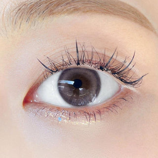 Close-up shot of model's eye adorned with Chuu Milk & Tea Cream Grey color contact lenses with prescription, complemented by minimalist eye makeup, showing the brightening and enlarging effect of the circle contact lens on dark brown eyes.