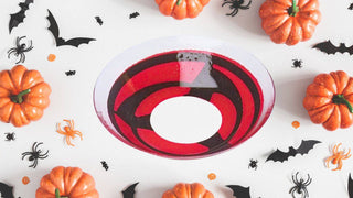 Red and black spiral contact lens surrounded by mini pumpkins, plastic spiders, and paper bats on a white surface.