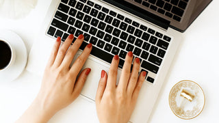 Hands with glossy terracotta red polished nails typing on a laptop keyboard on a white desk.