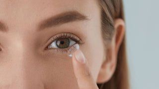 A woman holds a clear contact lens on her fingertip near her hazel eye.