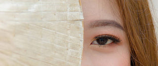 Close-up of a woman with a dark brown eye and warm eye makeup partially covered by a woven straw hat.