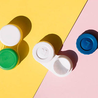 Contact lens cases and caps in green, white, and blue on a yellow and pink background.
