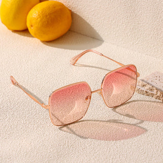 A pair of stylish EyeCandys Bermuda Oversized Square Sunglasses with Glacier and Pink Swizzle lenses and a gold frame is placed on a textured light-colored surface. Two yellow lemons are positioned in the background, casting soft shadows. The overall scene suggests a summery, sunny vibe.