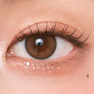 A close-up of a brown eye with long lashes, soft peach eyeshadow, and smooth skin, enhanced naturally by Chuu Aery Rose Lucid Brown (10pk) color contacts from Chuu. A beauty mark sits below the lower lash line.