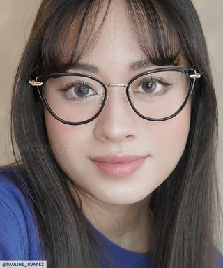 A person with long, straight dark hair and bangs is wearing EyeCandys Aura Cat Eye glasses and a blue top, looking at the camera with a slight smile.