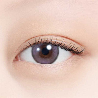 A close-up of a human eye with long lashes wearing N's Collection Blueberry Jam Violet (10pk) colored contact lens. The fair, smooth skin is brightly lit, highlighting the soft violet hue from N's Collection.