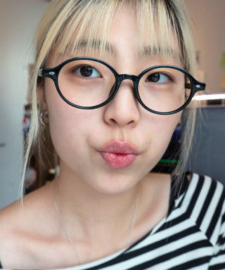 A person with blonde hair and a striped shirt wears EyeCandys Chase Round Oval glasses, looking at the camera with pursed lips under soft indoor lighting.