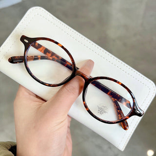 A hand holds a pair of EyeCandys Chase Round Oval Glasses with clear lenses, their round tortoiseshell frames resting on a white case, set against a smooth grayish background.